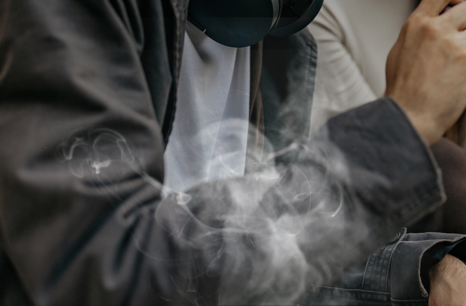 사람이 검은색 재킷을 입고 흰색 셔츠를 입고 있으며, 손에는 무언가를 쥐고 있습니다. 주변에는 연기가 피어오르고 있습니다.