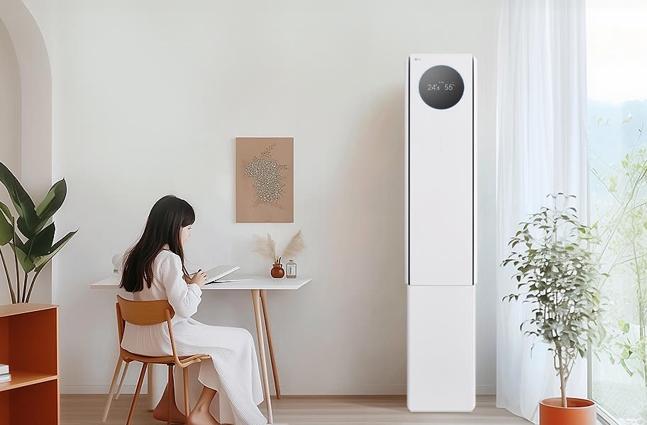 에어컨 상단에는 원형 디스플레이가 있으며, 24.5°와 55%라는 숫자가 표시되어 있습니다. 왼쪽에는 흰색 테이블과 나무 의자에 아이가 앉아 있으며, 테이블 위에는 책과 작은 장식품이 있습니다. 벽에는 식물 액자가 걸려 있으며, 오른쪽에는 커튼이 드리워진 창문과 화분이 보입니다.