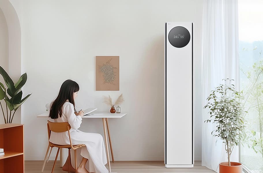 에어컨 상단에는 원형 디스플레이가 있으며, 24.5°와 55%라는 숫자가 표시되어 있습니다. 왼쪽에는 흰색 테이블과 나무 의자에 아이가 앉아 있으며, 테이블 위에는 책과 작은 장식품이 있습니다. 벽에는 식물 액자가 걸려 있으며, 오른쪽에는 커튼이 드리워진 창문과 화분이 보입니다.