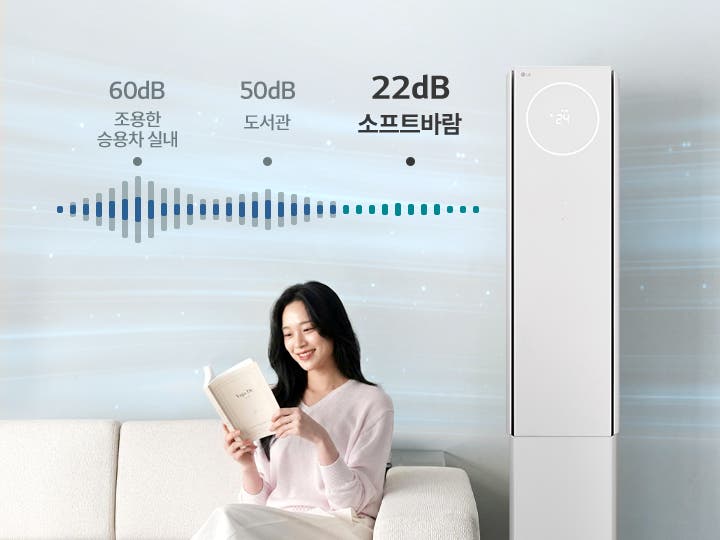 "해당 이미지는 에어컨의 소프트바람 기능에 대해 설명하는 이미지로 저소음 냉방 기능과 사용 환경을 시각적으로 보여주고 있습니다. 우측에 스탠드 에어컨이 배치되어 있으며, 여성 모델이 베이지색 소파에 앉아 책을 읽고 있습니다. 에어컨 상단의 원형 디지털 디스플레이에는 온도 정보가 표시되어 있습니다. 이미지 상단에는 소음 비교를 나타내는 그래픽과 함께 60dB 조용한 승용차 실내, 50dB 도서관, 22dB 뷰I 에어컨 이라는 문구가 순서대로 배치되어 있습니다."