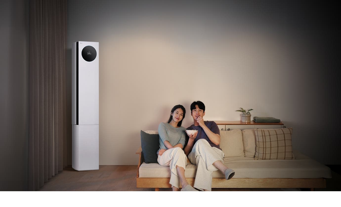 LG 휘센 AI 오브제컬렉션 뷰I 프로 에어컨이 왼쪽에 세워져 있으며, 원형 디스플레이에는 24.5℃라는 숫자가 표시되어 있습니다. 오른쪽에는 소파에 앉아 있는 두 사람이 보이며, 남성은 팝콘을 들고 있습니다. 소파 뒤쪽에는 수납장이 놓여 있고 그 위에는 식물과 수건이 놓여 있습니다.