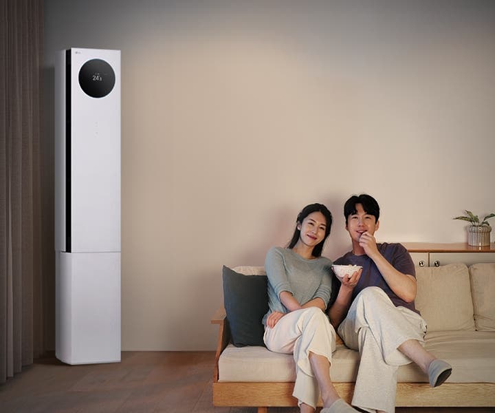 LG 휘센 AI 오브제컬렉션 뷰I 프로 에어컨이 왼쪽에 세워져 있으며, 원형 디스플레이에는 24.5℃라는 숫자가 표시되어 있습니다. 오른쪽에는 소파에 앉아 있는 두 사람이 보이며, 남성은 팝콘을 들고 있습니다. 소파 뒤쪽에는 수납장이 놓여 있고 그 위에는 식물과 수건이 놓여 있습니다.
