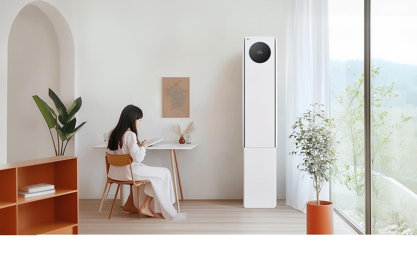 에어컨 상단에는 원형 디스플레이가 있으며, 24.5℃라는 숫자가 표시되어 있습니다. 왼쪽에는 흰색 테이블과 나무 의자에 아이가 앉아 있으며, 테이블 위에는 책과 작은 장식품이 있습니다. 벽에는 식물 액자가 걸려 있으며, 오른쪽에는 커튼이 드리워진 창문과 화분이 보입니다.