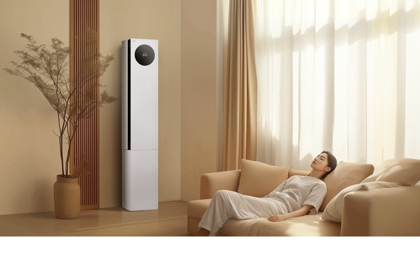 에어컨 상단에는 원형 디스플레이가 있으며, 24.5℃라는 숫자가 표시되어 있습니다. 에어컨 옆에는 나무 소재의 장식 패널과 화분이 놓여 있으며, 오른쪽에는 밝은 색상의 커튼이 드리워진 창문이 있습니다. 창문 앞에는 베이지색 소파가 놓여 있고, 소파에 한 여성이 편안하게 누워 있습니다. 실내는 따뜻한 색조로 꾸며져 있으며, 자연광이 부드럽게 들어오고 있습니다.