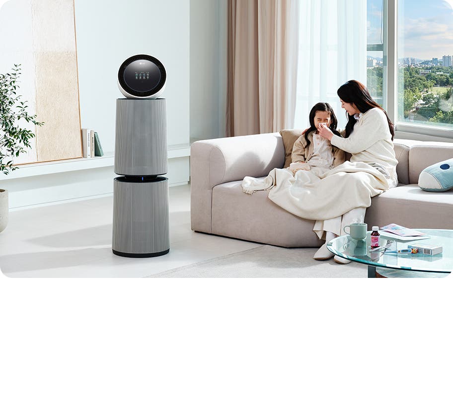 거실 공간에 LG 퓨리케어 AI 오브제컬렉션 360˚ 공기청정기가 놓여 있습니다. 소파 위에는 두 사람이 담요를 덮고 앉아 있으며, 한 여성이 아이의 코를 풀어주고 있는 모습입니다. 테이블 위에는 컵, 약봉투, 약이 놓여 있습니다.
