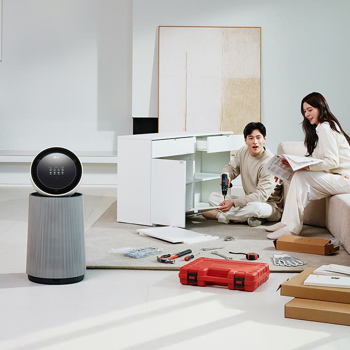 거실 공간에 LG 퓨리케어 AI 오브제컬렉션 360˚ 공기청정기가 놓여 있습니다. 가구를 조립하고 있는 두 사람의 모습이 보입니다. 주변에는 조립 중인 서랍장과 공구가 있습니다.