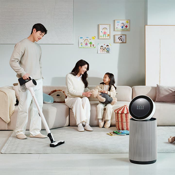 거실 공간에 LG 퓨리케어 AI 오브제컬렉션 360˚ 공기청정기가 있습니다. 소파에 앉아 있는 남성과 뛰어노는 아이, 그리고 청소기를 들고 있는 여성이 보입니다. 벽에는 그림이 걸려 있고, 바닥에는 화분과 장난감 등이 배치되어 있습니다.