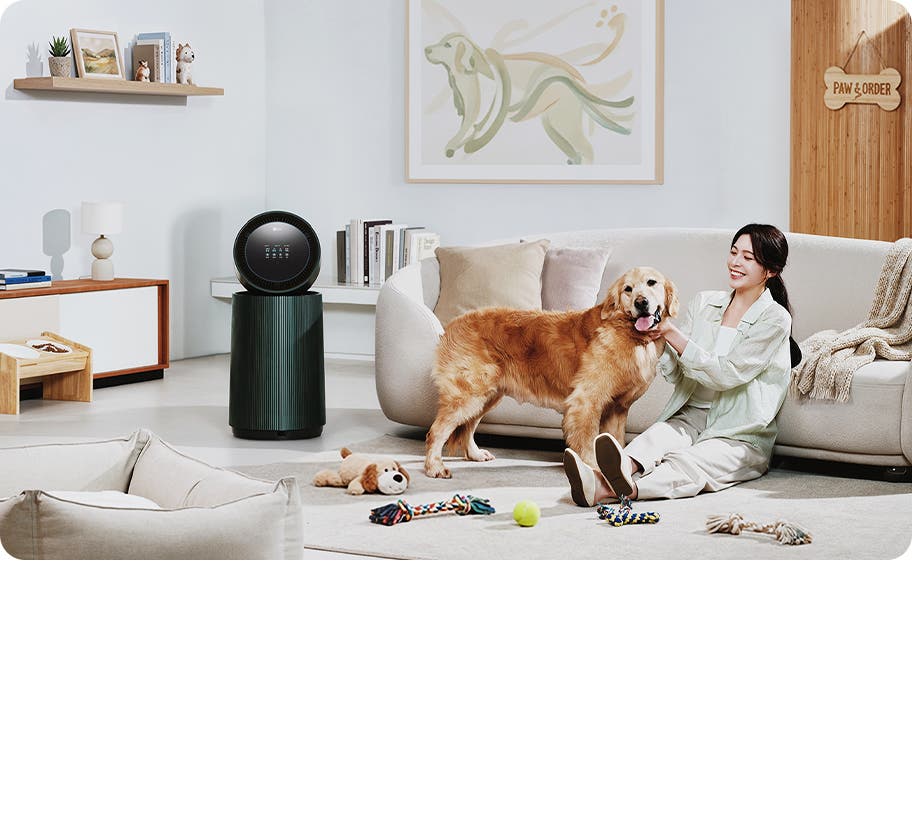 거실 공간에 LG 퓨리케어 AI 오브제컬렉션 360˚ 공기청정기가 놓여 있습니다. 한 여성이 앉아있는 강아지를 만지고 있으며 주변에는 강아지 장난감이 있습니다. 소파가 보이며, 벽에는 강아지 그림이 걸려 있고 선반에는 책과 장식품들이 놓여 있습니다.