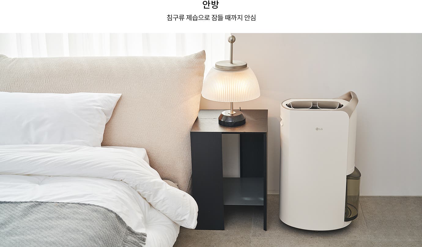 침대와 베개가 있는 침실의 모습입니다. 침대 옆에는 작은 테이블과 램프가 놓여 있으며, 그 옆에는 LG 로고가 있는 제습기가 있습니다. 텍스트로는 안방 침구류 제습으로 잠들 때까지 안심이라고 적혀 있습니다.