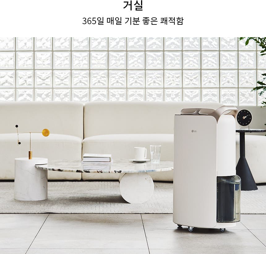 거실 공간에 흰색 소파와 대리석 테이블이 놓여 있으며, 테이블 위에는 책과 컵이 있습니다. 소파 뒤에는 유리 블록 벽이 있고, 한쪽에는 식물이 배치되어 있습니다. 바닥에는 카펫이 깔려 있으며, LG 로고가 있는 제습기가 있습니다. 상단에는 거실과 365일 매일 기분 좋은 쾌적함이라는 문구가 적혀 있습니다.