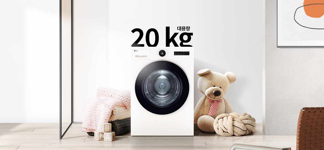 세탁기가 놓여 있으며, 상단에 대용량 20kg이라는 텍스트가 있습니다. 세탁기 주변에는 분홍색 니트 담요가 담긴 바구니와 곰 인형, 장난감 블록이 있습니다. 벽에는 추상적인 그림이 걸려 있습니다. 바닥은 나무 마루로 되어 있습니다.
