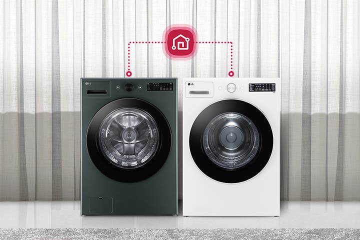 두 대의 LG 세탁기가 나란히 놓여 있으며, 각 세탁기 위에는 작은 디지털 패널이 있습니다. 세탁기 위쪽에는 스마트홈 아이콘이 연결된 모습이 보입니다. 배경에는 커튼이 드리워져 있고, 세탁기 옆에는 회색 의자와 둥근 테이블이 놓여 있습니다. 테이블 위에는 장식용 화병이 있습니다.