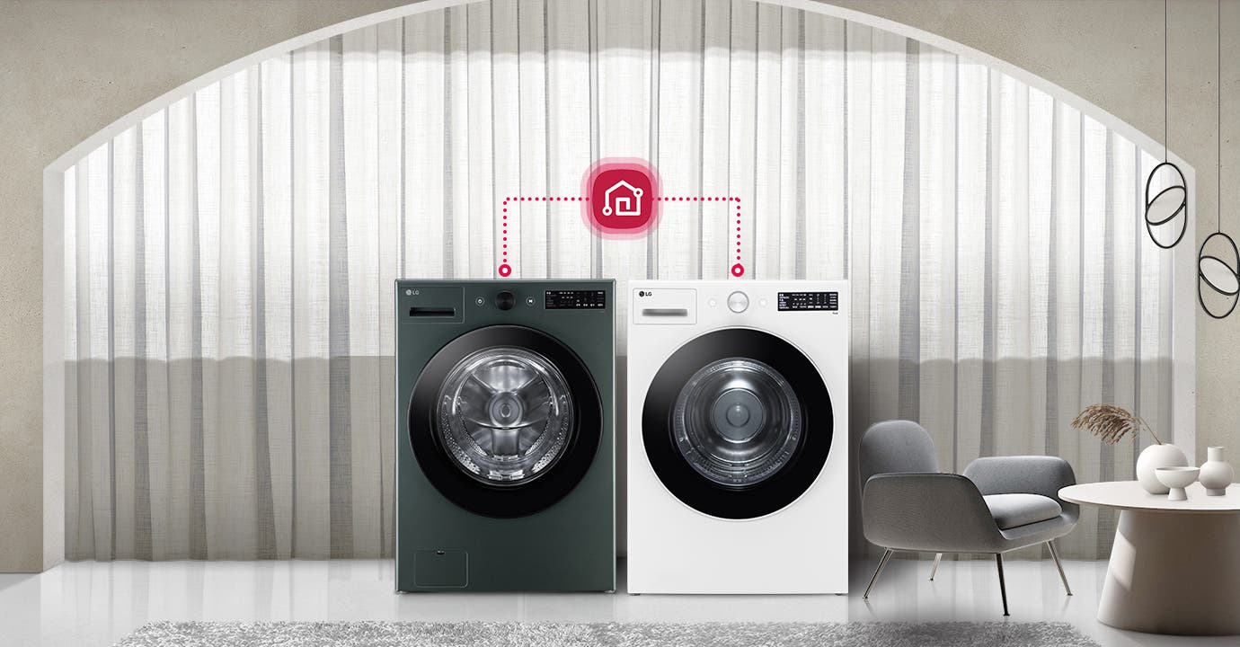 두 대의 LG 세탁기가 나란히 놓여 있으며, 각 세탁기 위에는 작은 디지털 패널이 있습니다. 세탁기 위쪽에는 스마트홈 아이콘이 연결된 모습이 보입니다. 배경에는 커튼이 드리워져 있고, 세탁기 옆에는 회색 의자와 둥근 테이블이 놓여 있습니다. 테이블 위에는 장식용 화병이 있습니다.