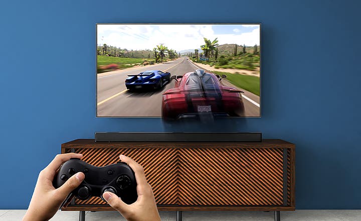벽에 걸린 TV 화면에 자동차 경주 게임이 표시되고 있으며, 두 대의 스포츠카가 도로를 달리고 있습니다. TV 아래에는 사운드바가 놓여 있고, 그 아래에는 나무 무늬의 장식장이 있습니다. 화면 앞쪽에는 게임 컨트롤러를 잡고 있는 손이 보입니다. 