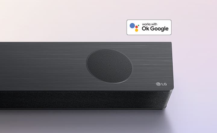 검은색 사운드바의 상단에 원형 스피커가 있으며, LG 로고가 표시되어 있습니다. 배경에는 works with Ok Google이라는 텍스트가 포함된 작은 배지가 있습니다.