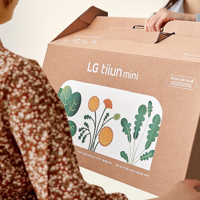 갈색 상자에 LG tiiun mini라는 글자가 인쇄되어 있으며, 상자에는 다양한 식물 그림이 그려져 있습니다. 상자 옆에는 Better Life for all이라는 문구가 적혀 있습니다. 두 사람이 상자를 들고 있는 모습입니다.