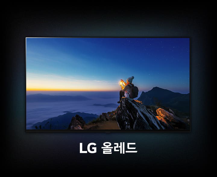 LG 올레드(OLED) TV 화면이 있습니다. TV 화면 속에는 어둠이 짙게 깔린 푸른 새벽 하늘 아래, 산 정상으로 보이는 바위 위에 한 사람이 앉아있는 풍경이 보입니다. 이 사람은 등산복을 입고 배낭을 메고 있으며, 두 손으로 밝게 빛나는 작은 랜턴을 감싸 쥐고 있습니다.  화면 아래에는 'LG 올레드'라는 흰색 텍스트가 중앙에 배치되어 있습니다.