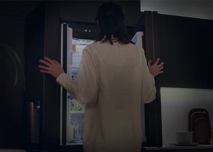 어두운 주방 공간에서 한 사람이 냉장고 문을 열고 있습니다. 냉장고 내부에는 여러 개의 선반과 식품들이 보이며, 왼쪽 문에는 작은 수납 공간이 있습니다.