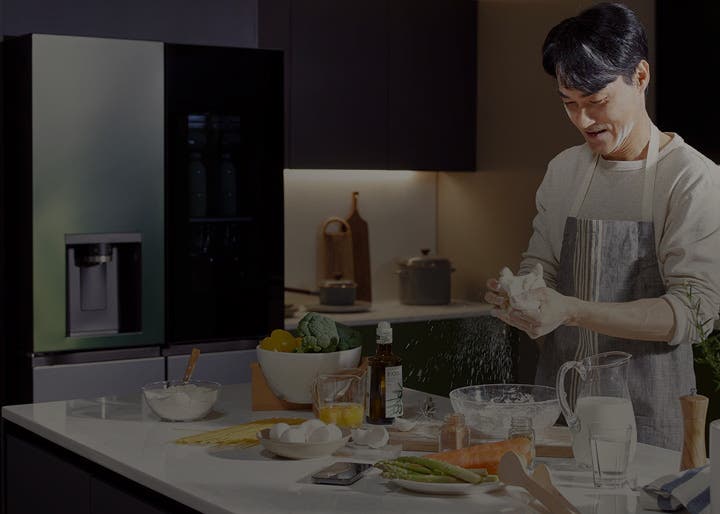 주방 공간에 있는 남성이 앞치마를 두르고 반죽을 하고 있으며, 그 옆에는 다양한 요리 재료가 놓여 있습니다. 왼쪽에는 냉장고가 보이며, 냉장고 문에는 물과 얼음을 받을 수 있는 디스펜서가 있습니다. 주방 조리대 위에는 계란, 밀가루, 아스파라거스, 브로콜리, 오일 병, 우유가 담긴 유리잔 등이 놓여 있습니다.