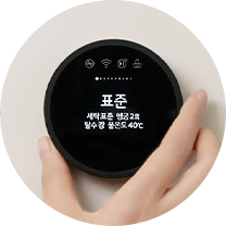 검은색 원형 디스플레이에 표준이라는 글자가 중앙에 표시되어 있으며, 그 아래로 세탁표준, 행굼 2회, 탈수 강, 물온도 40°C라는 텍스트가 보입니다. 디스플레이는 손으로 조작할 수 있도록 기기에 부착되어 있으며, 한 손이 디스플레이를 잡고 있는 모습이 보입니다.