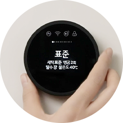 검은색 원형 디스플레이에 표준이라는 글자가 중앙에 표시되어 있으며, 그 아래로 세탁표준, 행굼 2회, 탈수 강, 물온도 40°C라는 텍스트가 보입니다. 디스플레이는 손으로 조작할 수 있도록 기기에 부착되어 있으며, 한 손이 디스플레이를 잡고 있는 모습이 보입니다.
