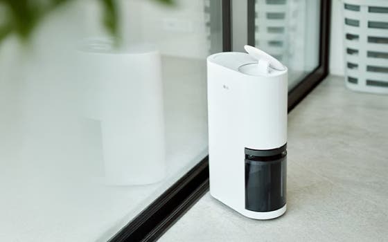 LG 제습기가 밝은 회색 타일 바닥 위에 놓여 있습니다. 제습기는 매끄러운 흰색 외관을 지녔으며, 상단에는 사용자 인터페이스와 조작용 덮개가 있습니다. 하단에는 투명한 검정색 물탱크가 자리하고 있습니다. 제습기는 유리문 옆에 위치해 있으며, 문과 제습기 표면에는 은은한 반사가 비칩니다.