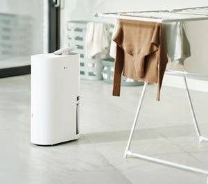 밝은 회색 타일 바닥에 놓인 흰색 LG 제습기, 뒤쪽에는 흰색 옷걸이에 갈색 스웨터와 젖은 옷들이 걸려 있고, 흐릿한 배경에는 플라스틱 세탁 바구니가 보입니다.