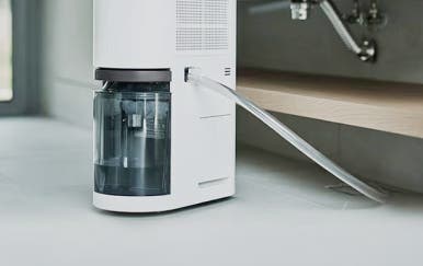 흰색 제습기 뒷면에 호스가 연결되어 부엌 싱크대 배수관 아래로 이어져 있습니다.
