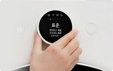 흰색 가전 제품의 검정색 원형 디스플레이를 손으로 조작하고 있으며, 화면에는 ‘표준’이라는 글자와 함께 절약 모드, 자동, 건조 정도 표준, 건조 시간 등 다양한 설정이 표시되어 있습니다.