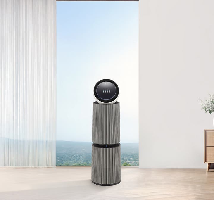 밝은 대리석 바닥에 LG 퓨리케어 AI 오브제컬렉션 360˚ 공기청정기가 놓여 있으며, 배경에는 흰색 벽과 창문이 있고, 창밖으로는 흐릿한 녹색 풍경이 보입니다. 공기청정기는 원통형 디자인으로 하단과 중간 부분이 세로 줄무늬가 있는 회색톤이며, 상단은 흰색 원형 패널과 검은색 테두리, 작은 디스플레이 화면으로 구성되어 있습니다.