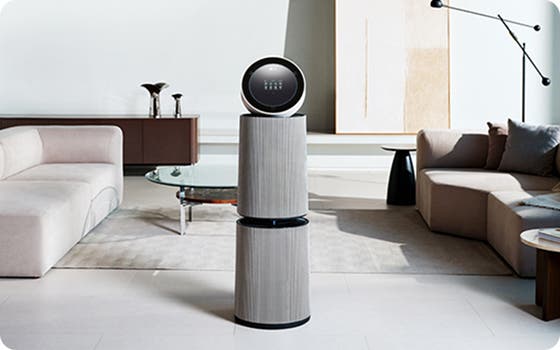 거실 공간에 LG 퓨리케어 AI 오브제컬렉션 360˚ 공기청정기가 놓여 있습니다. 양쪽에 베이지색 소파가 있으며, 쿠션과 담요가 놓여 있습니다. 유리 상판의 원형 테이블이 있고 그림과 검은색 스탠드 조명이 보입니다.