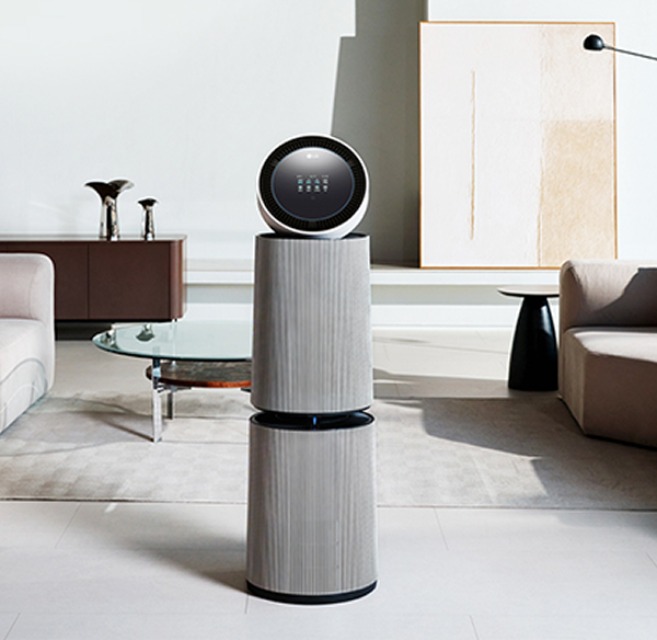 거실 공간에 LG 퓨리케어 AI 오브제컬렉션 360˚ 공기청정기가 놓여 있습니다. 양쪽에 베이지색 소파가 있으며, 쿠션과 담요가 놓여 있습니다. 유리 상판의 원형 테이블이 있고 그림과 검은색 스탠드 조명이 보입니다.