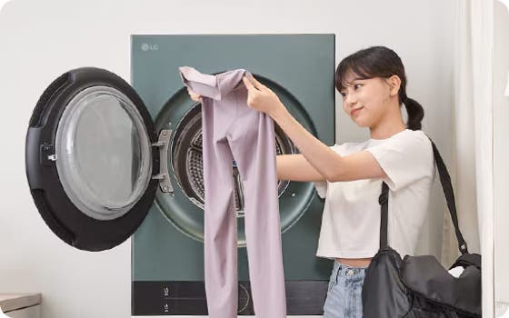 집에서 문이 열린 LG 건조기 앞에 선 여성이 라벤더색 레깅스를 넣으려 하고 있습니다. 그녀는 검은색 가방을 메고 있으며 미소를 짓고 있습니다.