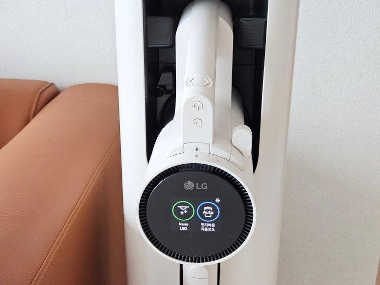 코드제로 A9S가 충전 거치대에 세워져 있으며, 디스플레이 화면에 Nano LED와 먼지비움 자동모드가 표시됨. 왼쪽에는 갈색 가죽 소파가 있음.