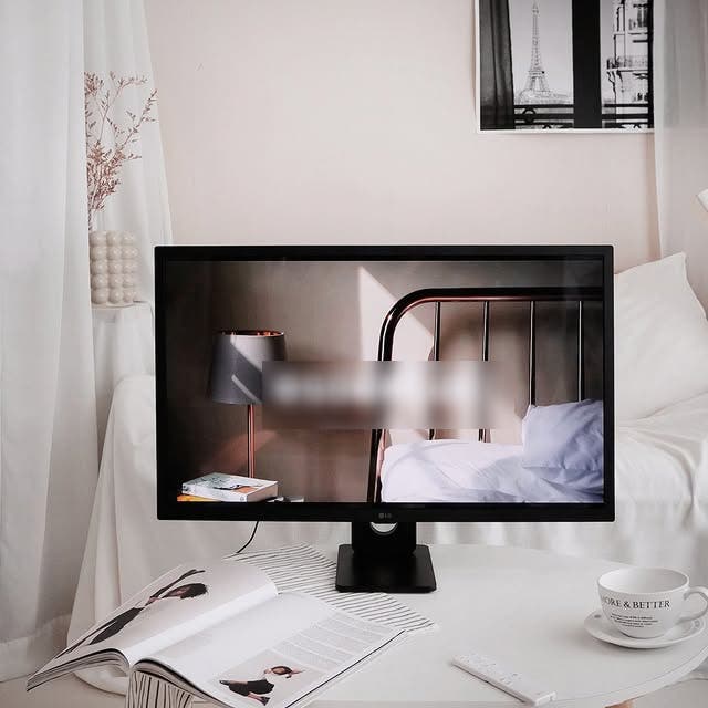 LG 스마트모니터가 책상 위에 놓여 있으며, 화면에는 모던한 침실 장면이 표시되어 있다. 책상이 있는 공간은 아늑한 인테리어로 꾸며져 있으며, 커피잔과 잡지가 함께 놓여 있어 편안한 분위기를 연출한다.