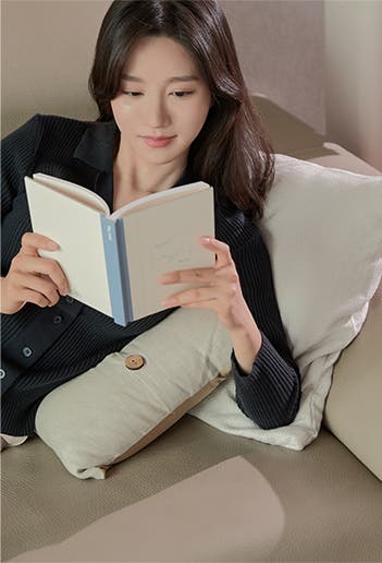 소파에 기대어 앉아 책을 읽는 여성. 편안한 자세로 독서에 집중하며 여유로운 시간을 보내는 모습.