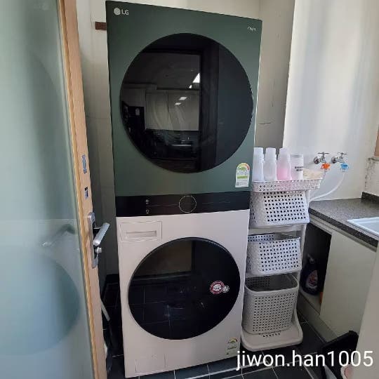 베란다 공간에 세탁기와 건조기가 세로로 배치되어 있고, 옆에는 세탁 용품을 보관하는 선반이 정리되어 있으며, 하얀색 커튼이 걸려 있음