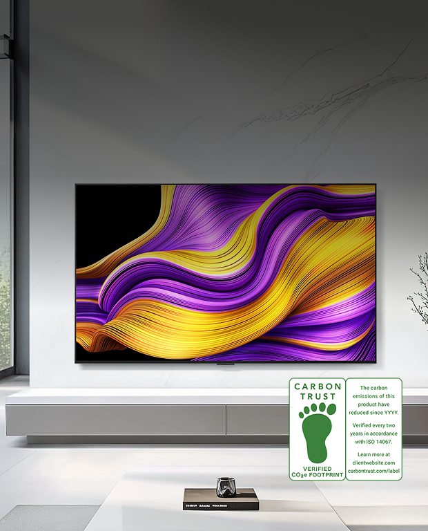 LG OLED TV 제품 이미지입니다. 흰색 벽면에 LG OLED TV가 벽걸이로 설치되어 있으며, 화면에는 보라색과 노란색의 웨이브 형태 추상 그래픽이 표시되어 있습니다. 우측 하단에 CARBON TRUST VERIFIED CO₂e FOOTPRINT 인증 마크와 함께 'The carbon emissions of this product have reduced since YYYY. Verified every two years in accordance with ISO 14067. Learn more at carbontrust.com/label' 텍스트가 표시되어 있습니다.
