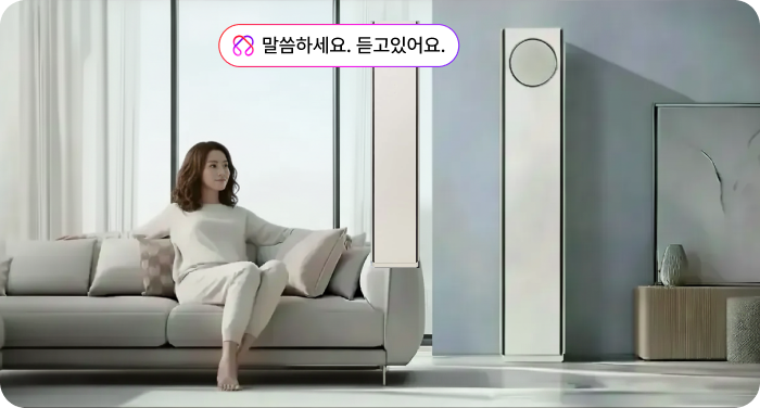 거실 소파에 편안하게 앉아 있는 여성이 슬림한 디자인의 LG 스탠드 에어컨을 바라보는 모습. 에어컨 상단에는 '말씀하세요. 듣고있어요.'라는 문구와 함께 음성 인식 기능을 나타내는 대화창 아이콘이 떠 있음