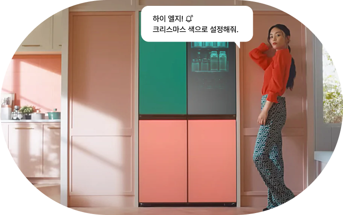 LG 냉장고 AI 음성 명령 기능 이미지입니다. 여성이 상단 그린, 하단 핑크 컬러의 LG 냉장고 옆에 서 있으며, 음성 명령 말풍선이 표시됩니다. 명령: '하이 엘지! 크리스마스 색으로 설정해줘.'