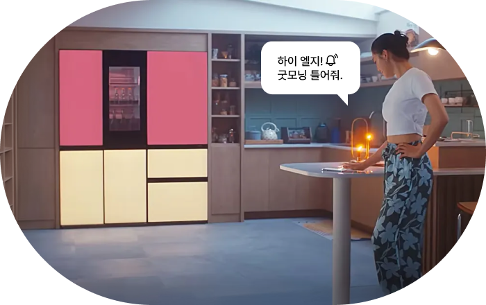 LG 냉장고 AI 음성 명령 기능 이미지입니다. 여성이 주방에서 상단 핑크, 하단 베이지 컬러의 LG 냉장고가 있는 공간에 서 있으며, 음성 명령 말풍선이 표시됩니다. 명령: '하이 엘지! 굿모닝 틀어줘.'