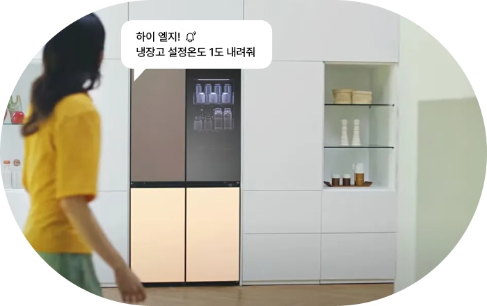 LG 냉장고 AI 음성 명령 기능 이미지입니다. 여성이 상단 브라운, 하단 베이지 컬러의 LG 냉장고 쪽으로 걸어가는 모습과 함께 음성 명령 말풍선이 표시됩니다. 명령: '하이 엘지! 냉장고 설정 온도 1도 내려줘.'