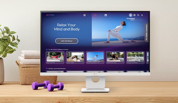 화이트 모니터 화면에는 'Relax Your Mind and Body'라는 문구와 함께 요가 동작을 하는 영상이 재생 중입니다. 하단에는 'Try Alo Moves Wellness Content' 섹션에 다양한 요가 및 명상 프로그램 썸네일들이 나열되어 있으며, 책상 위에는 보라색 덤벨과 수건이 놓여 있어 홈트레이닝 환경을 연출하고 있습니다.