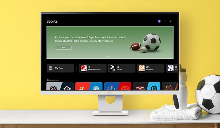 노란색 배경의 화이트 모니터 화면에 'Sports' 메뉴가 실행 중입니다. 'Register your favourite team/player' 문구와 축구공, 미식축구공 이미지가 보이며, 하단에는 다양한 스포츠 팀의 로고와 라이브 경기 정보가 나열되어 있습니다. 책상 위에는 축구공과 물병이 놓여 있어 활기찬 스포츠 테마를 강조합니다.