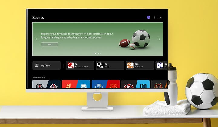 노란색 배경의 화이트 모니터 화면에 'Sports' 메뉴가 실행 중입니다. 'Register your favourite team/player' 문구와 축구공, 미식축구공 이미지가 보이며, 하단에는 다양한 스포츠 팀의 로고와 라이브 경기 정보가 나열되어 있습니다. 책상 위에는 축구공과 물병이 놓여 있어 활기찬 스포츠 테마를 강조합니다.